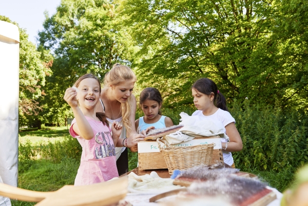 Eine junge Frau steht zwischen drei kleinen Kindern an einem Tisch im Freien. Sie zeigt zwei Kindern das Filzen, ein Kind zeigt etwas in die Kamera und lacht.