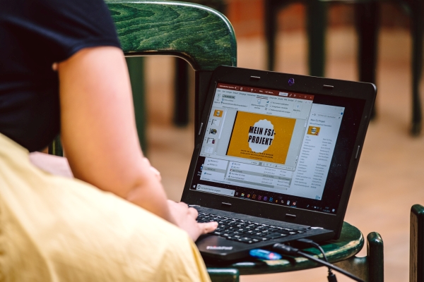 Eine junge Frau bedient einen Laptop, der auf einem Stuhl steht. Auf dem Laptop sieht man eine Präsentation mit dem Titel "Mein FSJ Projekt"
