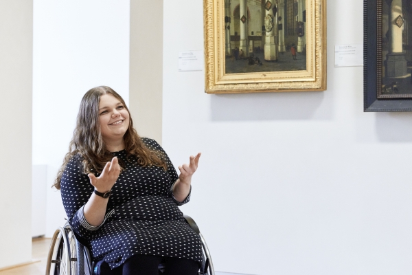 Eine Frau, die im Rollstuhl sitzt, gibt eine Erklärung zu einem Gemälde.