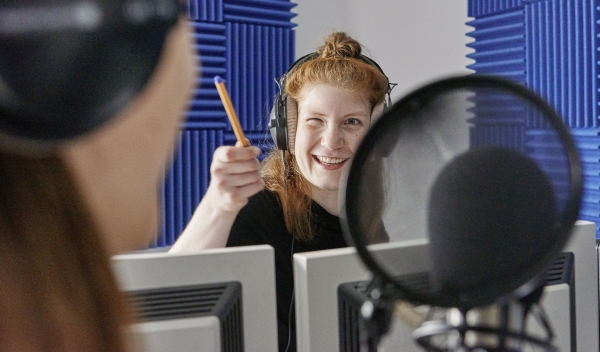Eine junge Frau steht hinter Computerbildschirmen in einem Studio mit Mikrofonen. Sie zwinkert und zeigt mit einem Stift auf eine andere Person.