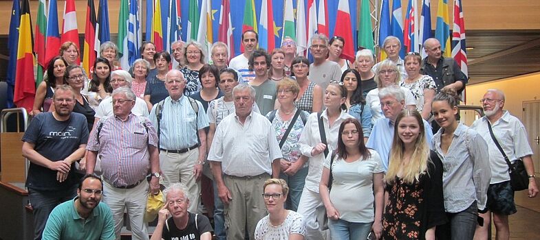 Eine Gruppe von älteren und jüngeren Menschen steht vor einem Wals aus Flaggen der Länder Europas. 