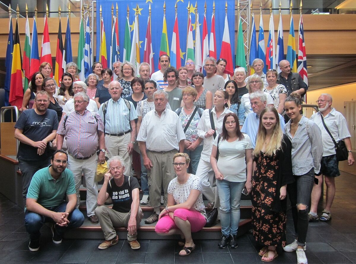 Eine Gruppe von älteren und jüngeren Menschen steht vor einem Wals aus Flaggen der Länder Europas.