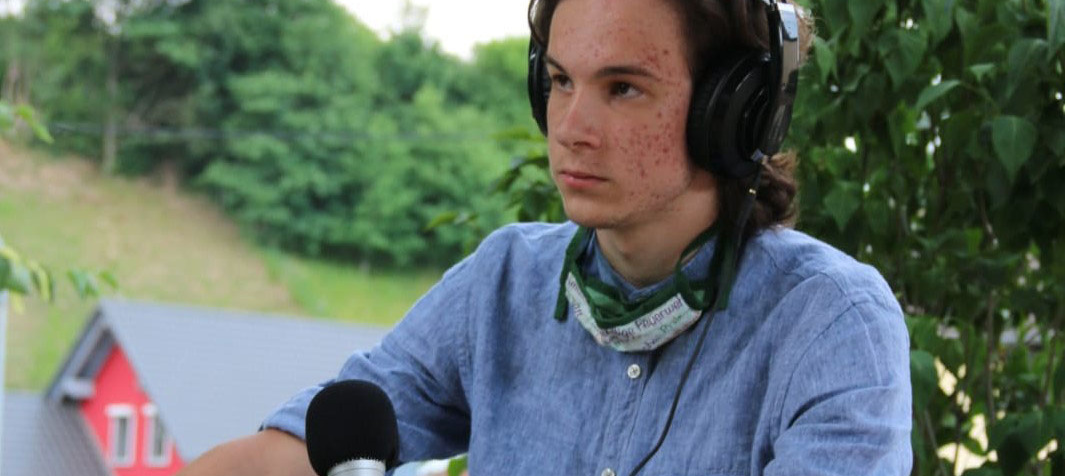 Ein Junge* sitzt auf einem Stuhjl. Er hat Kopfhörer auf und ein Mikrofon in der Hand.