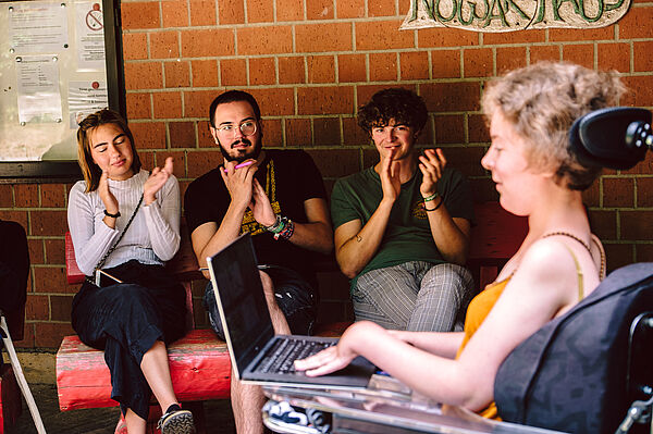 Sarah bei der Präsentation ihres eigenen Projekts Drei Personen sitzen an eine Hauswand gelehnt und klatschen Beifall. Sarah ist im Vordergrund zu sehen. Sie beendet gerade die Präsentation ihres eigenen Projekts.