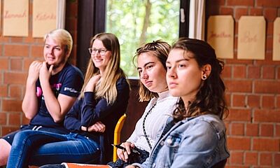 Vier junge Menschen sitzen in einer Reihe in einem Raum und blicken in eine Richtung.