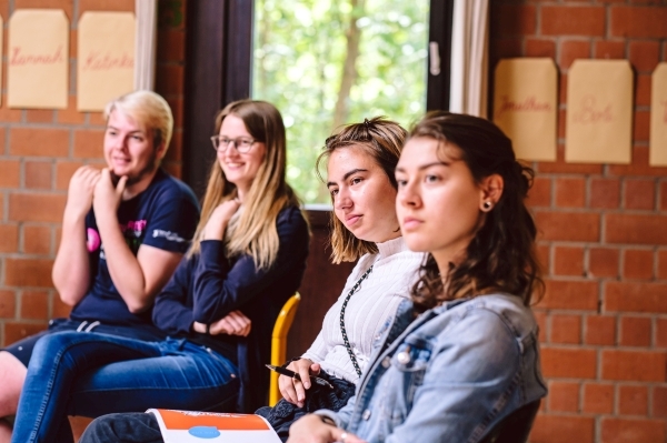 Vier junge Menschen sitzen in einer Reihe in einem Raum und blicken in eine Richtung.