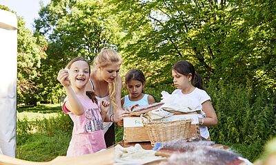 Eine junge Frau steht zwischen drei kleinen Kindern an einem Tisch im Freien. Sie zeigt zwei Kindern das Filzen, ein Kind zeigt etwas in die Kamera und lacht.