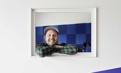 Ein junger Mann mit Basecap und grünschwarz kariertem Hemd schaut lächelnd aus einem Fenster. 
