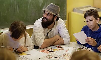 Ein junger Mann of Colour mit Hut sitzt an einem Tisch mit Kindern und bastelt.